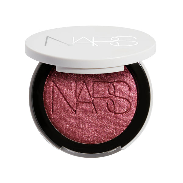 סומק פנינתי Light Reflecting Luminizing Blush של NARS, מוצג פתוח באריזה לבנה עגולה. הסומק בגוון ורוד-שזיפי עם גימור פנינתי וזוהר, המעניק ללחיים צבע עדין, ברק טבעי ומראה קורן.