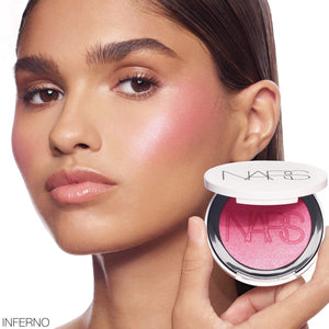 דוגמנית עונדת את הסומק הפנינתי Light Reflecting Luminizing Blush של NARS בגוון Inferno (Fuchsia), המעניק ללחיים צבע פוקסיה זוהר עם ברק פנינתי ומראה קורן. בתחתית התמונה מוצג הסומק באריזתו הלבנה הפתוחה.